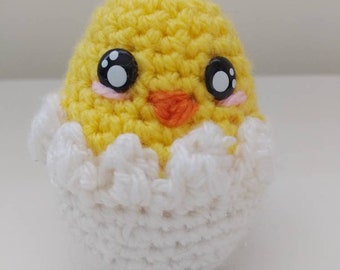 May include: A crocheted chick toy in a white eggshell. The chick has yellow yarn, black button eyes, and an orange beak.