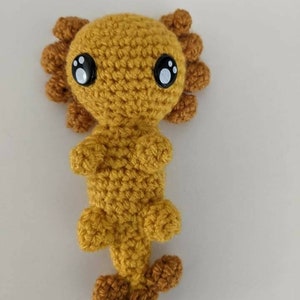 May include: A crocheted axolotl plush toy with large black eyes and a yellow body. The axolotl has a brown tail and brown frills around its head.