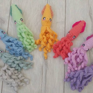 May include: A rainbow assortment of crocheted squid plushies. The squid are arranged in a semi-circle on a light wood surface. The squid are made in various colors, including green, yellow, orange, pink, purple, and gray.