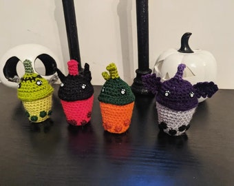May include: Four crocheted Halloween-themed boba tea cups. The cups are green, red, orange, and purple. Each cup has a different character on top, including a monster, a vampire, a ghost, and a bat.