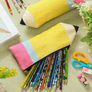 Puede incluir: Dos estuches de tela con forma de lápiz en amarillo, rosa y azul claro con puntas negras. Un estuche está abierto y muestra una colección de lápices con varios patrones. El diseño imita un lápiz clásico con una goma de borrar rosa.