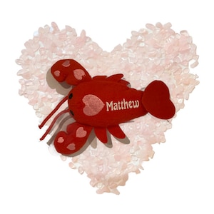 Puede incluir: Juguete de peluche de langosta roja con corazones rosas bordados. La langosta tiene el nombre "Matthew" bordado en su cuerpo. La langosta está sentada en una cama en forma de corazón de cristales de cuarzo rosa.