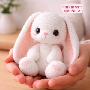 May include: A white crochet bunny with long ears, black eyes, and a pink nose and cheeks. The bunny's inner ears and the soles of its feet are also pink. The text "FLOPPY THE WHITE BUNNY PATTERN" is in a pink rectangle.