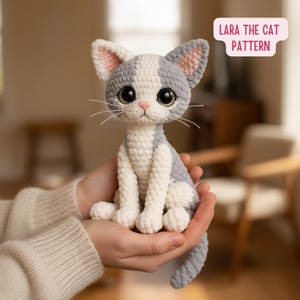Może przedstawiać: Ręcznie robiony szydełkowy kot w kolorze szarym, białym i różowym, z dużymi czarnymi oczami i wąsami. Kot trzymany jest w otwartych dłoniach. Tekst "Lara the Cat Pattern" znajduje się na różowo-białym banerze.