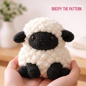 Puede incluir: Un peluche de oveja hecho a mano con un cuerpo blanco y esponjoso y una cara, orejas y pezuñas negras. El texto "SHEEPY THE PATTERN" se muestra en rosa. La oveja está sostenida en las manos de una persona.