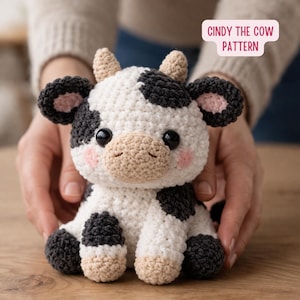 Può includere: Un peluche di mucca all'uncinetto, con macchie bianche e nere, occhi neri e guance rosa. La mucca ha muso, orecchie e corna beige. Il testo "CINDY THE COW PATTERN" è in un rettangolo rosa.