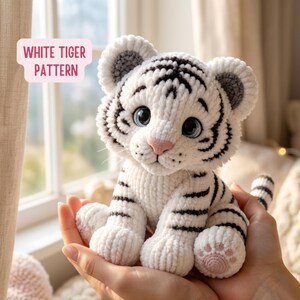 Puede incluir: Un peluche de tigre blanco hecho a mano con rayas negras, ojos azules y una nariz y almohadillas rosas. El texto "WHITE TIGER PATTERN" está en un cuadro rosa. El tigre está sentado y la imagen está bien iluminada.