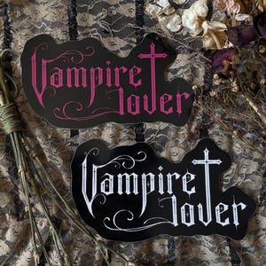 May include: Two black stickers with the words "Vampire Lover" in a gothic font. One sticker has pink text and a cross, the other has white text and a cross. The stickers are on a lace background.