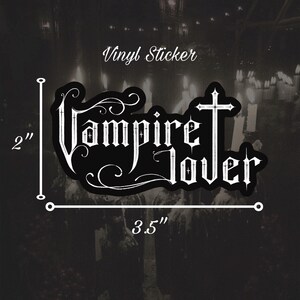 May include: Black vinyl sticker, 3.5 inches wide and 2 inches tall, with the words "Vampire Lover" in a gothic font. The sticker has a cross design and decorative swirls. The text "Vinyl Sticker" is above the main text.