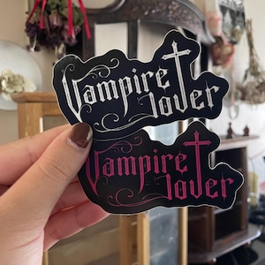 May include: Two black stickers with the words "Vampire Lover" in a gothic font, topped with a cross. One sticker has white text, the other has pink text. The stickers are held up in front of a blurred background.