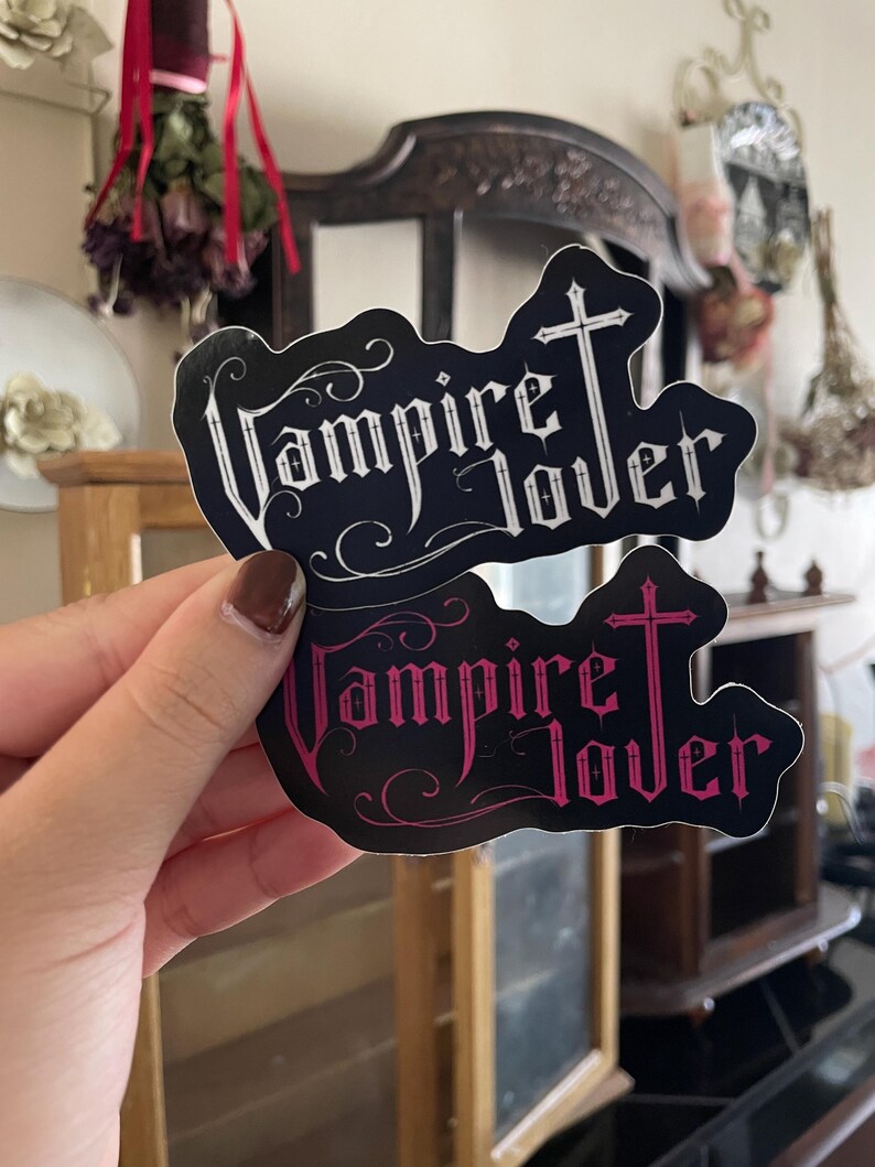 May include: Two black stickers with the words "Vampire Lover" in a gothic font, topped with a cross. One sticker has white text, the other has pink text. The stickers are held up in front of a blurred background.