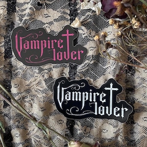 May include: Two stickers with the text "Vampire Lover". One sticker has pink text on a black background, and the other has white text on a black background. The stickers are in a gothic style.