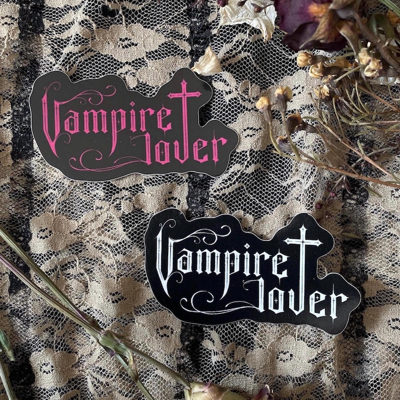 May include: Two stickers with the text "Vampire Lover". One sticker has pink text on a black background, and the other has white text on a black background. The stickers are in a gothic style.
