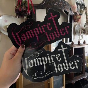 May include: Two black stickers with the words "Vampire Lover" in a gothic font, each with a cross design. One sticker has pink text, and the other has white text. The stickers are held up in front of a blurred background.