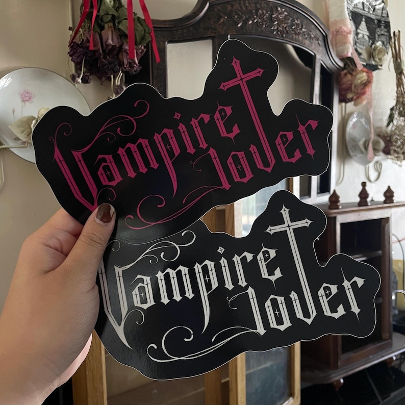 May include: Two black stickers with the words "Vampire Lover" in a gothic font, each with a cross design. One sticker has pink text, and the other has white text. The stickers are held up in front of a blurred background.