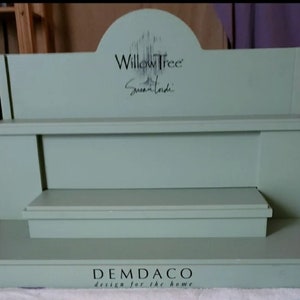 May include: A three-tiered wooden display stand painted in a light green color. The top tier features the text "Willow Tree" and "Susan Lordi" in black lettering. The bottom tier has the text "DEMDACO design for the home" in black lettering.