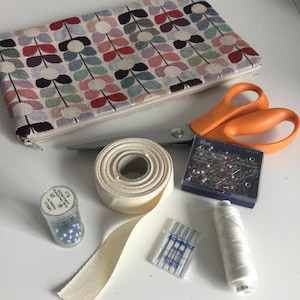 May include: A selection of sewing equipment including a floral patterned zip-up pouch, orange scissors, white thread, pins, needles, ribbon, and a small container of blue beads. The items are arranged on a white surface.