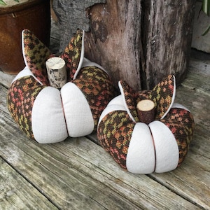 May include: Two fabric pumpkins featuring alternating panels of solid white canvas and a multicoloured woven pattern in shades of orange, brown, and green. Each pumpkin is topped with a small, cylindrical wooden stem.
