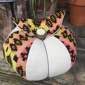 May include: A decorative pumpkin-shaped cushion featuring alternating panels of solid off-white fabric and a woven textile with a geometric pattern in yellow, orange, and black. A small, cylindrical wooden piece serves as the stem at the centre.