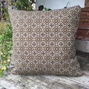 May include: A square throw cushion featuring a repeating geometric pattern in shades of brown and cream. The design consists of interconnected circular and square motifs woven into the fabric, creating a textured appearance across the entire surface.