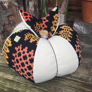 May include: A decorative fabric pumpkin featuring alternating panels of solid cream-coloured linen and black fabric with intricate geometric cross-stitch patterns in orange, yellow, and peach. The pumpkin is topped with a small, natural wooden stem.