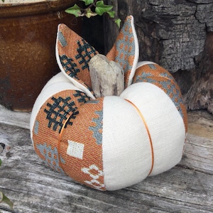 May include: A fabric pumpkin decoration featuring alternating panels of solid cream-coloured fabric and orange fabric with a dark grey geometric pattern. The pumpkin is topped with a central piece of light-coloured driftwood acting as a stem.