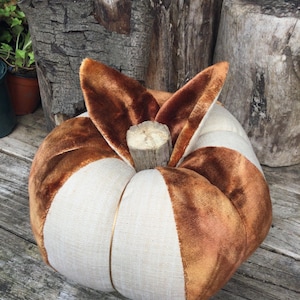 May include: A decorative pumpkin crafted from alternating panels of brown velvet and cream-coloured linen fabric. The pumpkin features a central stem made from a piece of natural, light-coloured wood, giving it a textured and organic appearance.