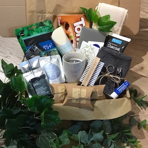 May include: A brown paper gift box filled with various self-care items. Includes a gray candle, a 2022 planner, a charcoal face mask, a nail brush, and a lip balm. Greenery and a box of chocolates are also visible.