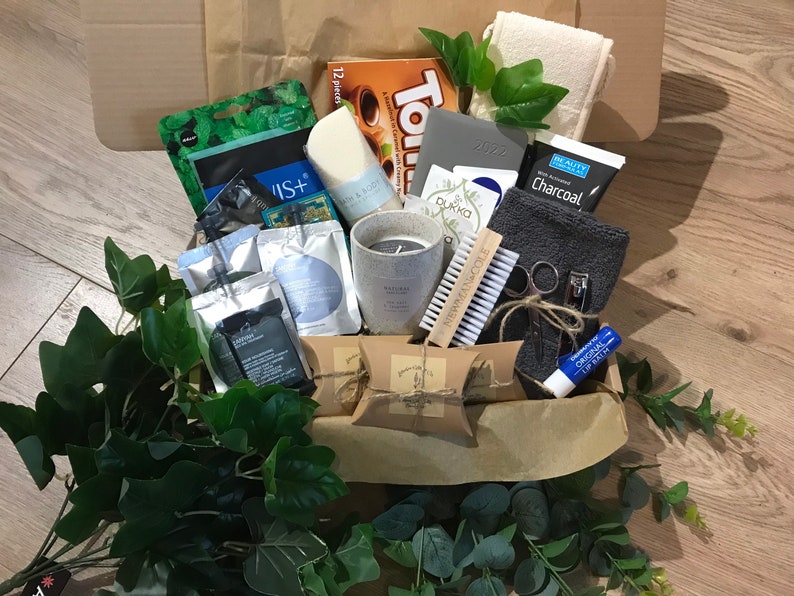 May include: A brown paper gift box filled with various self-care items. Includes a gray candle, a 2022 planner, a charcoal face mask, a nail brush, and a lip balm. Greenery and a box of chocolates are also visible.