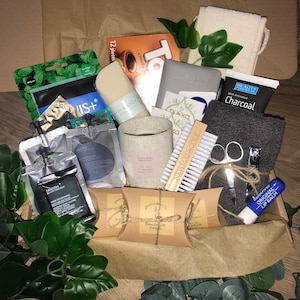 May include: A brown cardboard box filled with self-care products. Items include a candle, soap, charcoal face mask, nail clippers, lip balm, and a washcloth. Green foliage and brown paper fill the box.