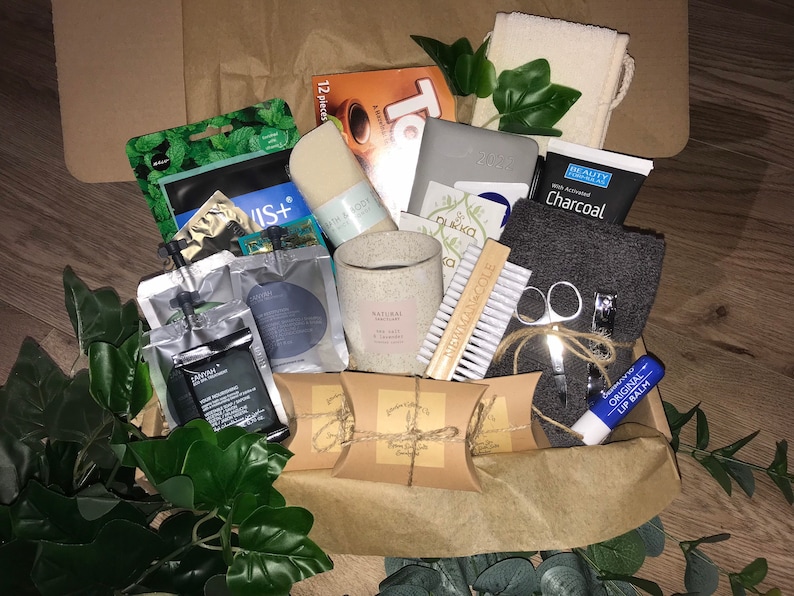 May include: A brown cardboard box filled with self-care products. Items include a candle, soap, charcoal face mask, nail clippers, lip balm, and a washcloth. Green foliage and brown paper fill the box.