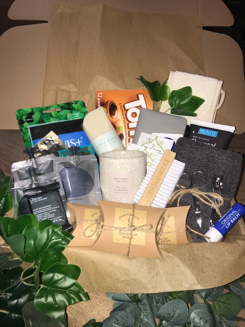 May include: A gift box filled with self-care items. Includes a candle, soap, lip balm, a 2022 planner, and other beauty products. The box is lined with brown paper and decorated with faux greenery.