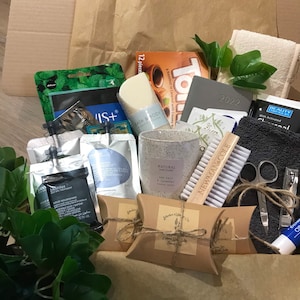 May include: A gift box filled with self-care items. Includes a candle, soap, face masks, a nail brush, nail clippers, a small towel, and other beauty products. The box is brown and the items are arranged inside.
