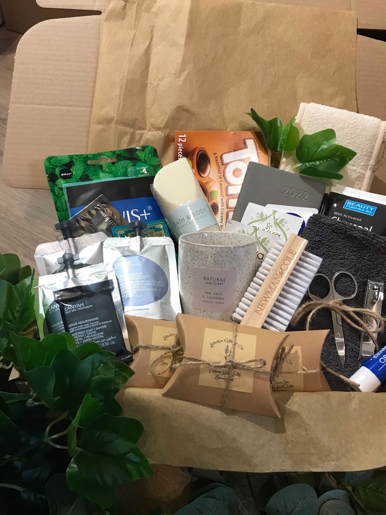 May include: A gift box filled with self-care items. Includes a candle, soap, face masks, a nail brush, nail clippers, a small towel, and other beauty products. The box is brown and the items are arranged inside.