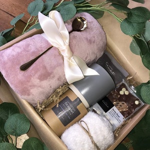 May include: A gift box filled with self-care items, including a pink plush blanket, a bath bomb, a white washcloth, a gray mug, and a chocolate bar. The box is lined with brown paper shreds.