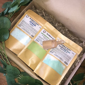 May include: Three brown paper bags with blue, green, and light blue bath salts inside. The bags are labeled "Bubble Dust" and have different scents: "Ocean", "Peppermint", and "Five for Him".