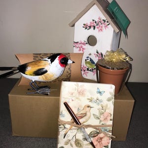 May include: A decorative bird figurine, a white birdhouse with pink floral details, a floral notebook with a pen, and a small terracotta pot with a gold bow. The notebook features the text "EUROPEAN ROBIN".