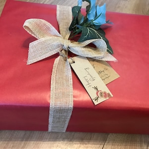 May include: A wrapped gift in red paper, adorned with a burlap ribbon tied in a bow. The bow is accented with faux greenery and a tag that reads "Happy Reading". A festive and decorative presentation.