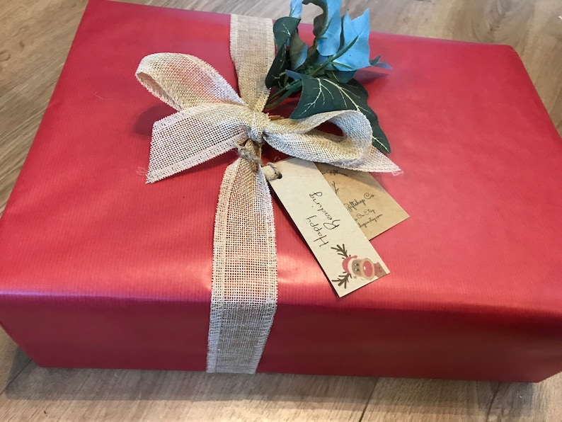 May include: A wrapped gift in red paper, adorned with a burlap ribbon tied in a bow. The bow is accented with faux greenery and a tag that reads "Happy Reading". A festive and decorative presentation.