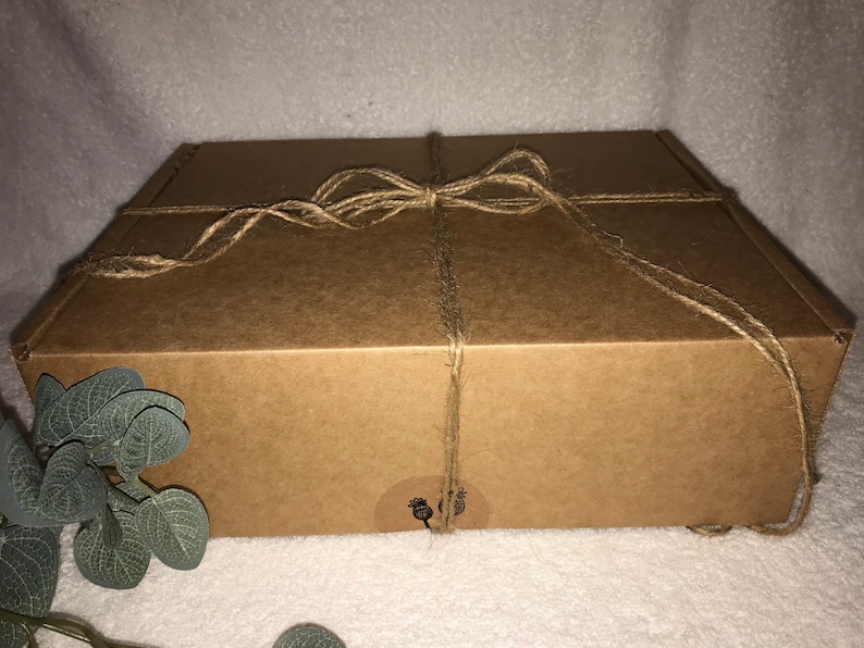 May include: A rectangular brown cardboard box tied with twine. The box has a small, round sticker on the front. The twine is wrapped around the box and tied in a bow. The box is sitting on a white surface.