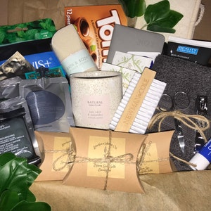 May include: A gift box filled with self-care items. Includes a candle, bath sponge, lip balm, nail clippers, a gray towel, and several small packages of beauty products. The box also contains a small notebook and a box of chocolates.