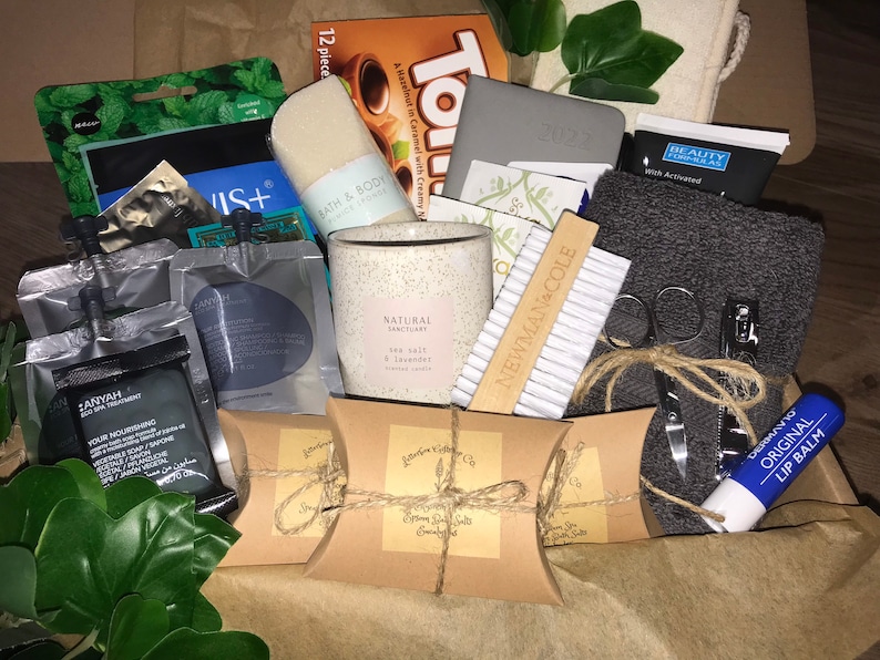May include: A gift box filled with self-care items. Includes a candle, bath sponge, lip balm, nail clippers, a gray towel, and several small packages of beauty products. The box also contains a small notebook and a box of chocolates.