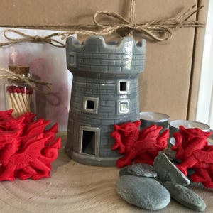 May include: A gray ceramic castle-shaped wax burner with a red dragon wax melt and a box of matches. The wax melt is shaped like a dragon and is red. The matches are in a glass jar with a cork top. The wax burner is on a wooden surface.