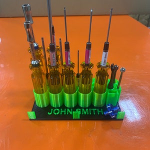 May include: A green 3D printed screwdriver holder with the name "JOHN SMITH" printed on the front. The holder has 10 slots for screwdrivers, and there are 9 screwdrivers in the holder. The screwdrivers are all different sizes and have different colored handles.