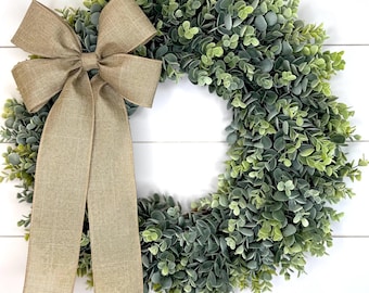 May include: A green eucalyptus wreath with a brown burlap bow. The wreath is made of artificial greenery and is perfect for adding a touch of nature to your home decor.