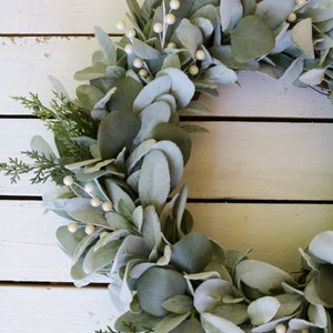 May include: A green and white eucalyptus wreath with white berries. The wreath is made of faux eucalyptus leaves and is perfect for a farmhouse or rustic decor style.