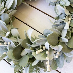 May include: A green and white faux eucalyptus wreath with white berries. The wreath is made of artificial leaves and berries and is perfect for decorating a home or office.