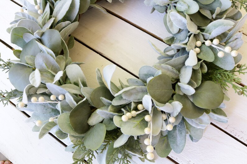 May include: A green and white faux eucalyptus wreath with white berries. The wreath is made of artificial leaves and berries and is perfect for decorating a home or office.