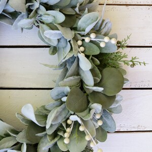 May include: A green and white faux eucalyptus wreath with white berries. The wreath is on a white wooden background.