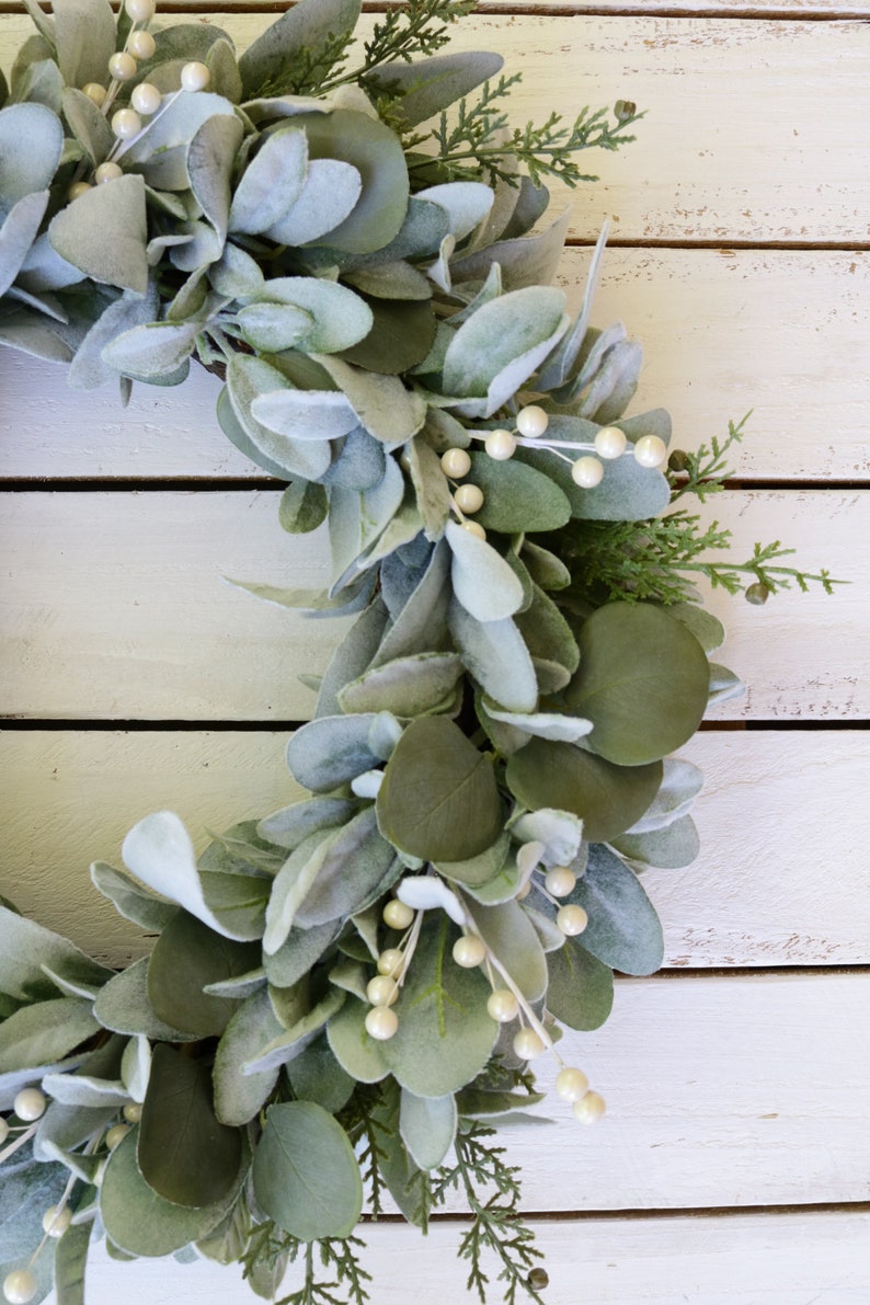 May include: A green and white faux eucalyptus wreath with white berries. The wreath is on a white wooden background.